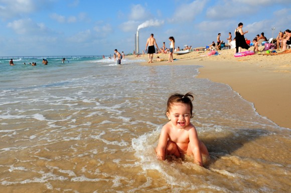 A baby playing in the surf on a beach with a polluting smoke stack in the background