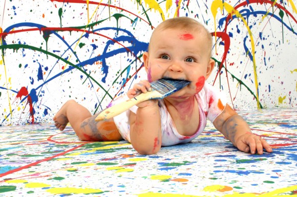 baby splattered with paint chewing on a paintbrush