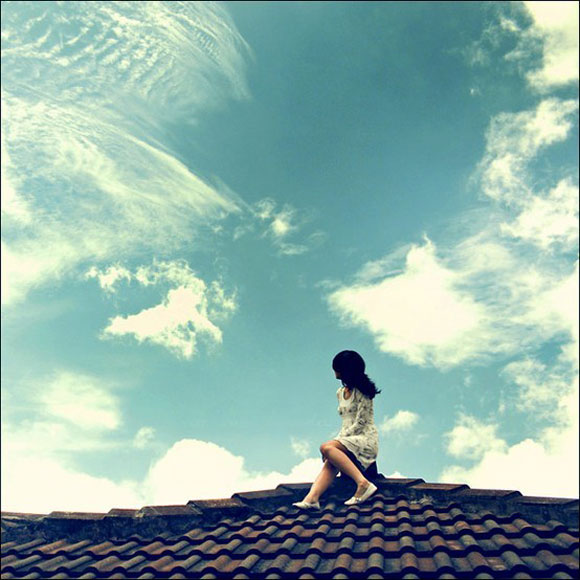 Woman sitting on roof with blue sky and clouds behind her