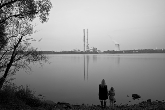 Mother and child hold hands, overlooking a lake and smokestacks